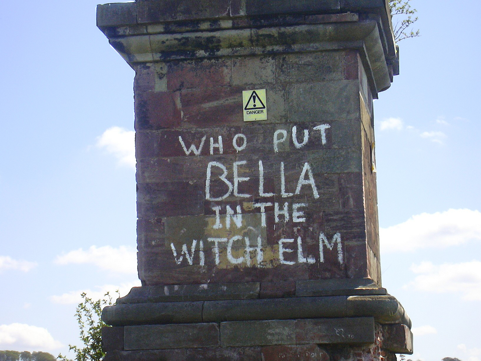 A weathered stone obelisk plinth with white lettering painted across it reading WHO PUT BELLA IN THE WITCH ELM, a yellow triangular danger sign fixed above the text, against a blue sky.